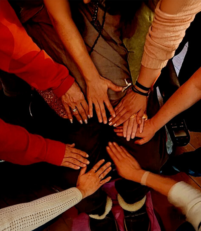 Mehrere Hände verschiedener Personen liegen unterstützend und gemeinsam auf den Beinen einer sitzenden Person und symbolisieren Zusammenhalt und Fürsorge