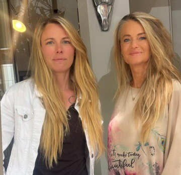 Zwei Frauen stehen nebeneinander in einem hellen Innenraum und blicken freundlich in die Kamera.