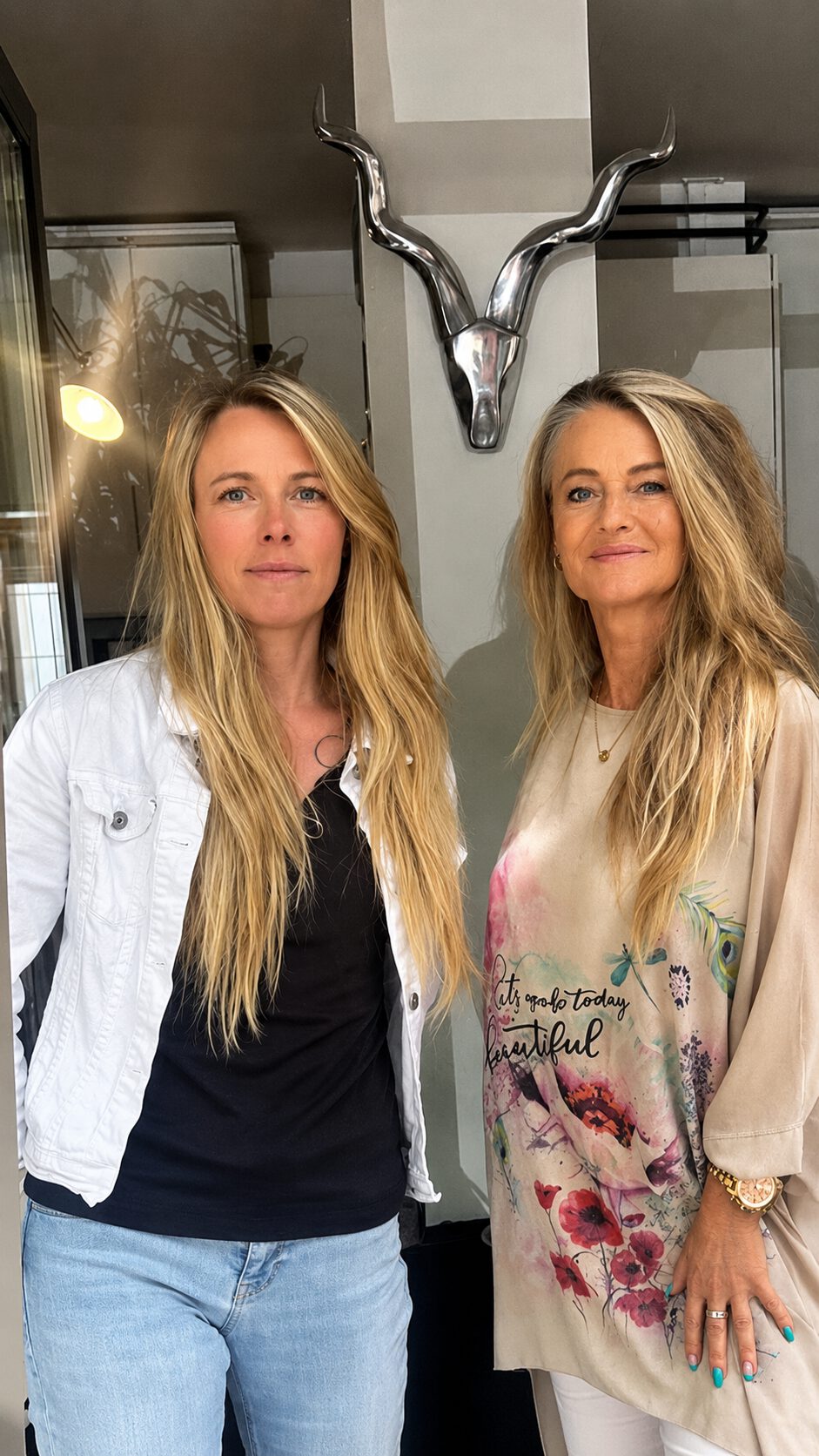 Zwei Frauen stehen nebeneinander in einem hellen Innenraum und blicken freundlich in die Kamera.